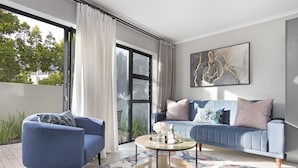 Una sala de estar moderna con un sofá azul, un sillón azul, una mesa de centro de vidrio y un ventanal con cortinas.