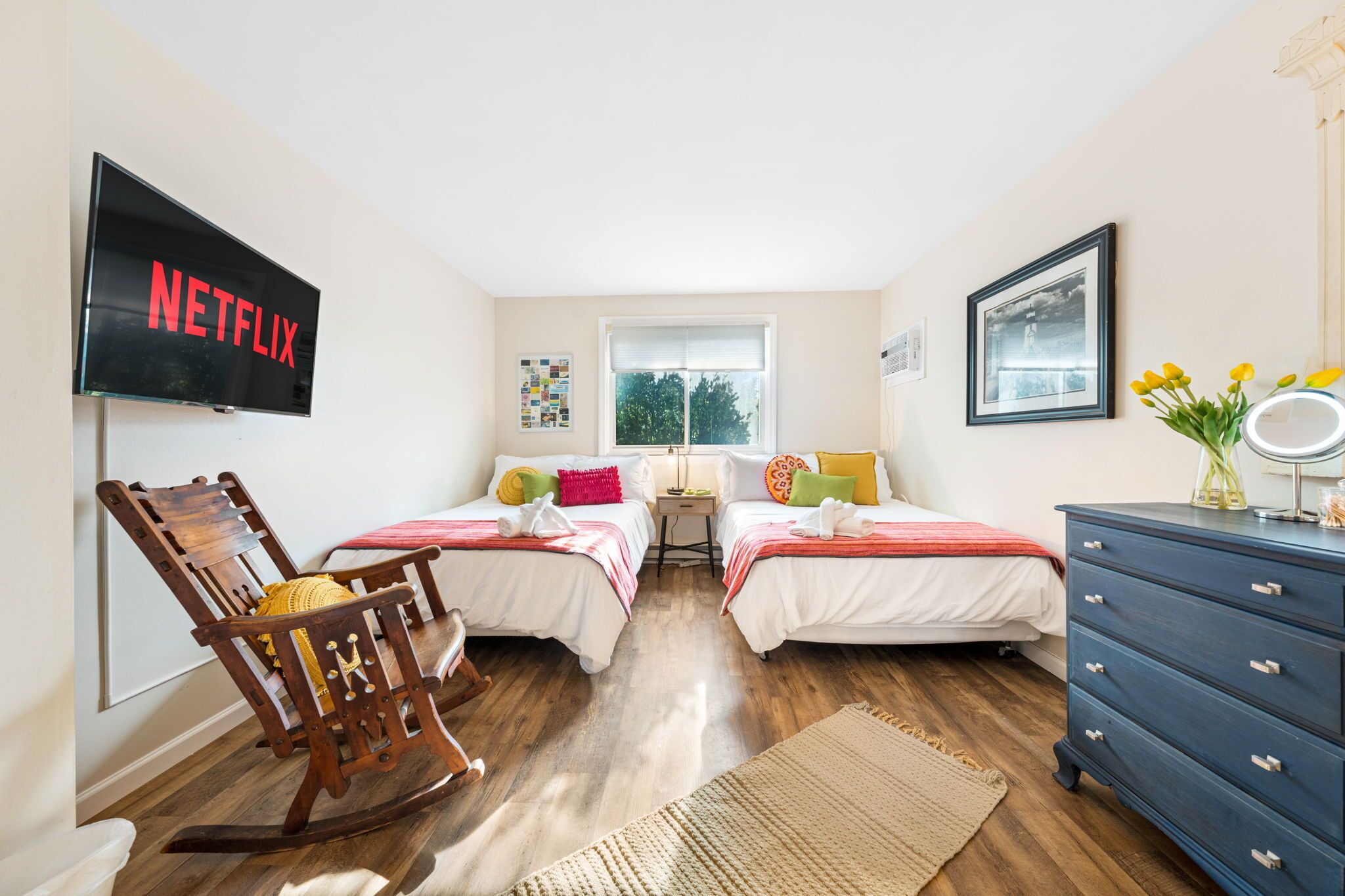 A bedroom with two beds, a rocking chair, a dresser, a TV, and a window with a view of trees.