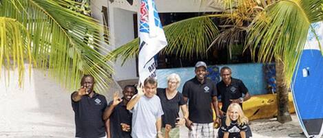 Un grupo de personas posando en una playa arenosa con palmeras y un edificio al fondo.