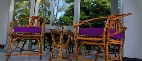 Two wicker chairs with purple cushions and a small round table with a decorative item, all set on a tiled floor under a glass roof with a view of trees and sky.