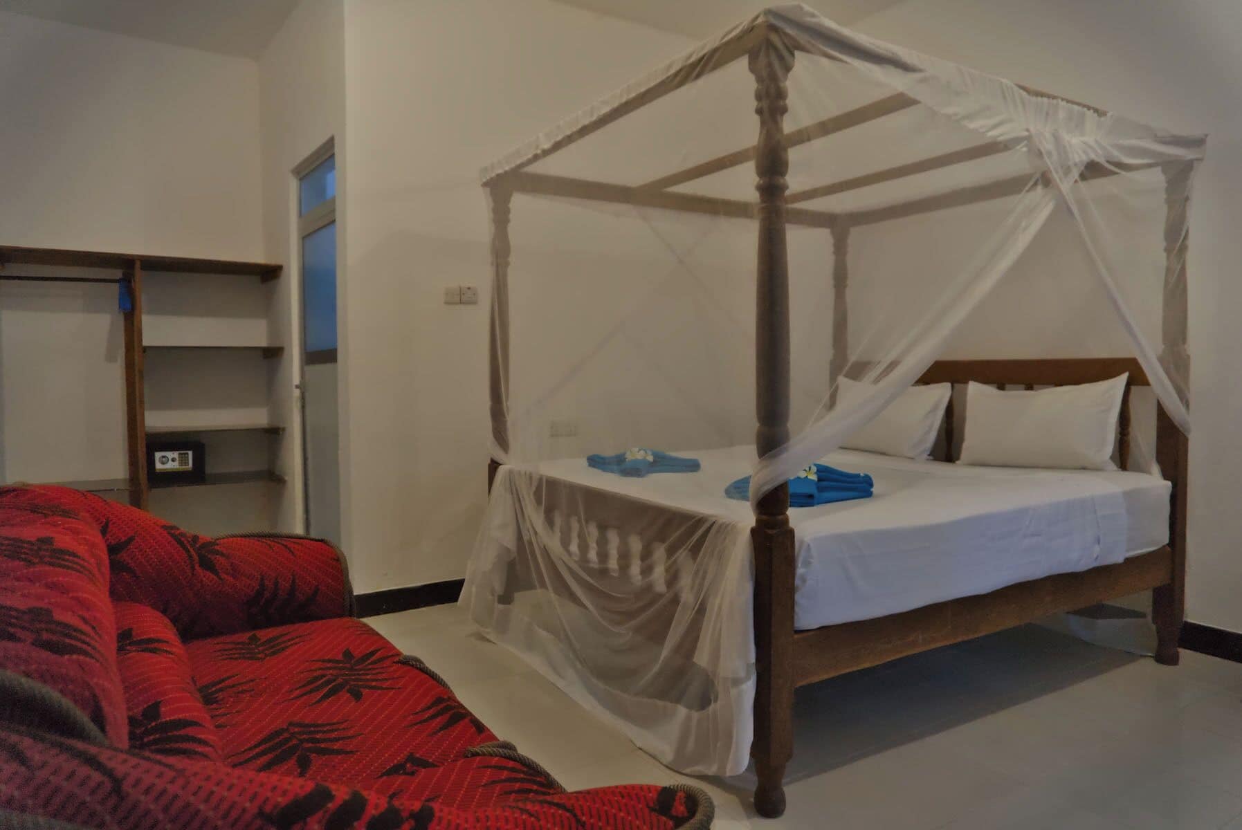 A four-poster canopy bed with a white bedspread and pillows, a red patterned sofa, and a wooden nightstand with a radio.