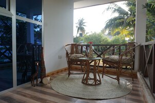 A dining area with wicker chairs and a round table, large windows with a view of palm trees, and a wooden floor.