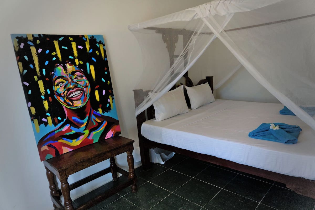A room with a bed under a white canopy, a painting on the wall, and a small wooden stool.