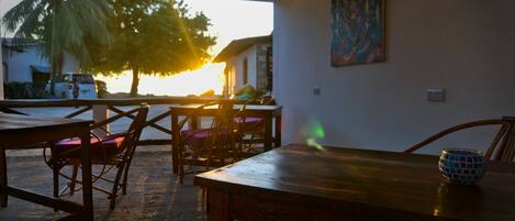 An indoor dining area with wooden tables and chairs, a view of the sunset through a window, and a painting on the wall.