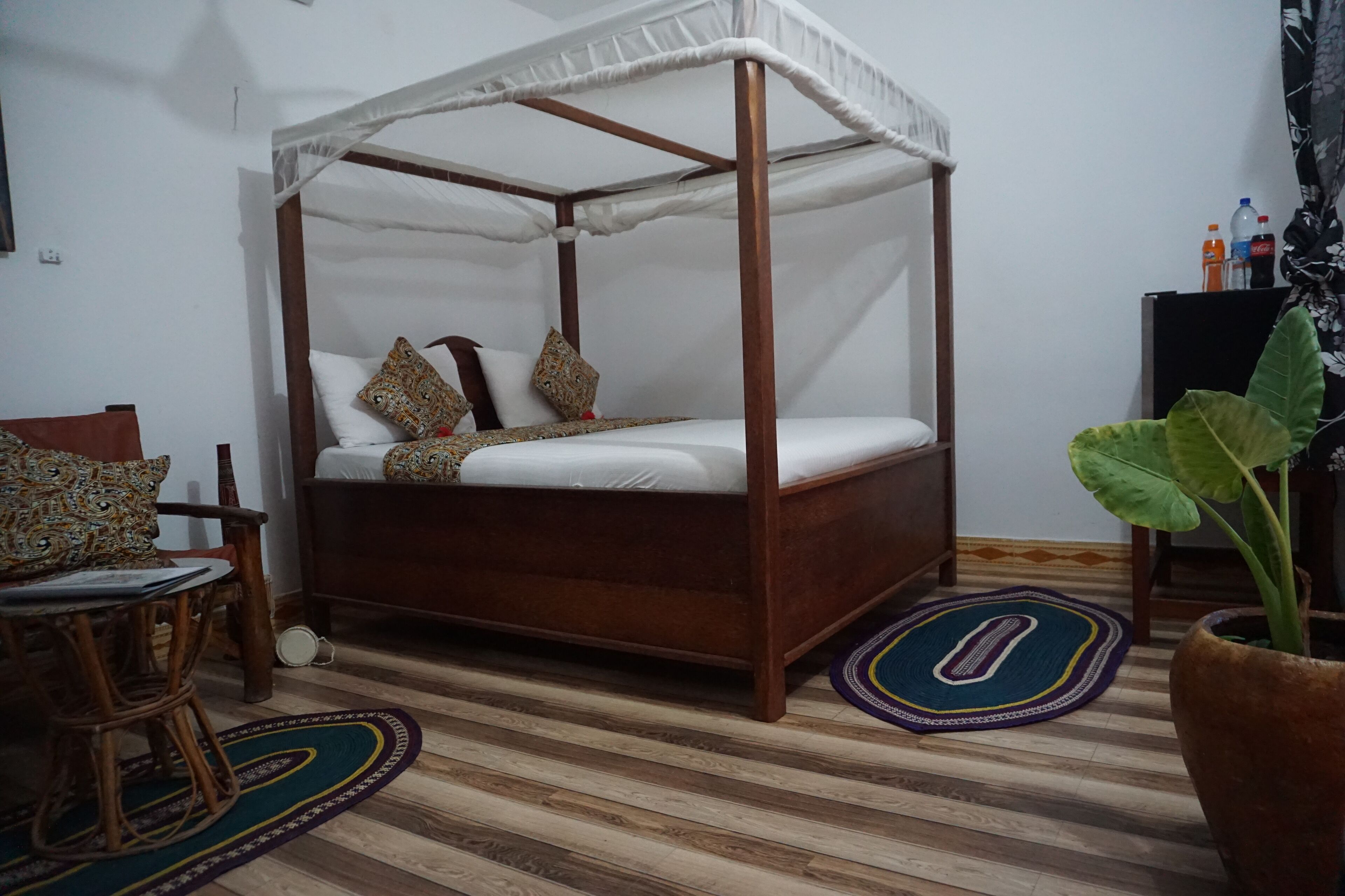 Un letto a baldacchino con tenda, un pavimento in legno, una sedia in rattan e una pianta in vaso.