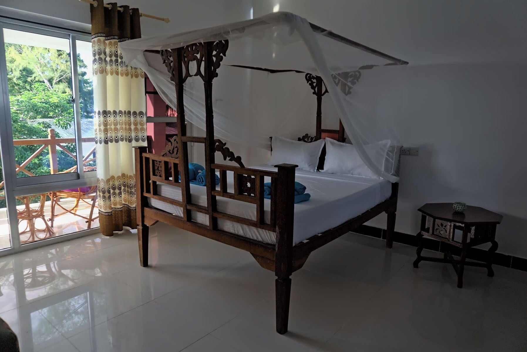 A four-poster bed with a canopy, a small table, and a view of greenery outside.