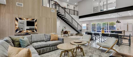 Un salón amplio con un sofá gris, una mesa redonda de centro y una escalera que sube a un piso superior.