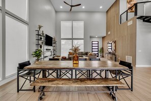 A spacious dining area with a large wooden table and chairs, a ceiling fan, and a modern kitchen in the background.