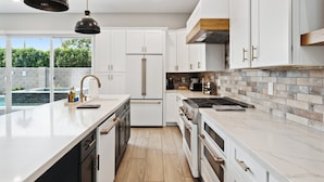 A modern kitchen with white cabinets, a central island, stainless steel appliances, and a view of a swimming pool. - Lavish Luxuries-Pool/Spa, Games, Walk To Old Town! (Scottsdale)