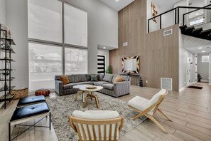 A spacious living room with a large window, a grey sectional sofa, a round coffee table, and a wooden chair. - Lavish Luxuries-Pool/Spa, Games, Walk To Old Town! (Scottsdale)