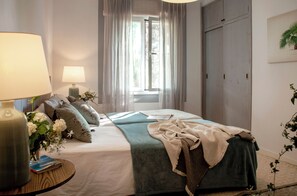 Une chambre à coucher bien rangée, avec un lit, des lampes de chevet, une table de chevet ornée d’un vase de fleurs et une fenêtre avec des rideaux.
