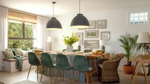 Un coin repas avec une table dressée pour quatre personnes, entourée de chaises vertes et d’un fauteuil en osier, avec une fenêtre et des rideaux en arrière-plan.