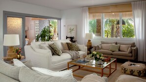 Un salon avec un canapé blanc, un canapé beige et une table basse. On y trouve deux lampes, une plante et une fenêtre avec des rideaux.