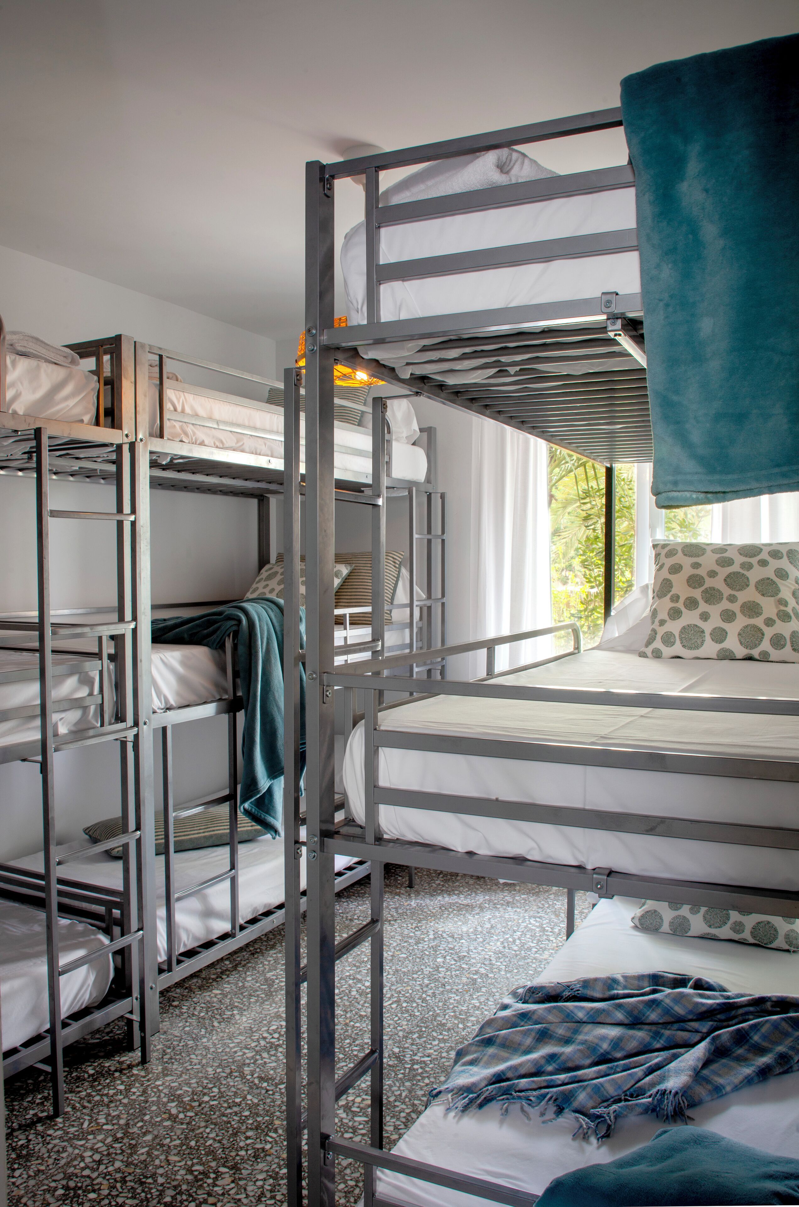 A bunk bed room with metal railings, white bedding, and a view of greenery outside.