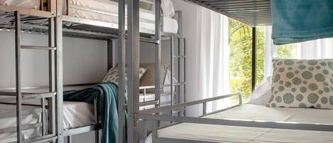 A bunk bed room with metal railings, white bedding, and a view of greenery outside.