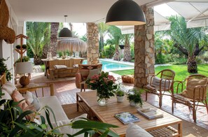 A sunlit patio with a pool, wicker furniture, and lush greenery.