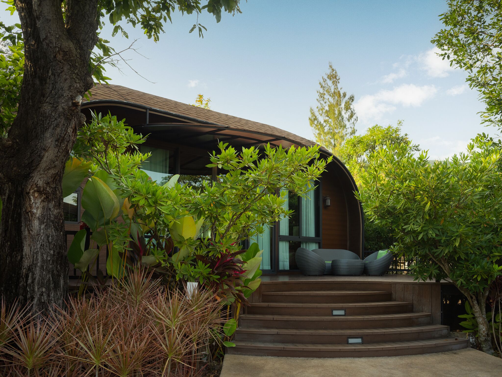 Una casa moderna con techo curvo, rodeada de exuberante vegetación y un gran árbol.