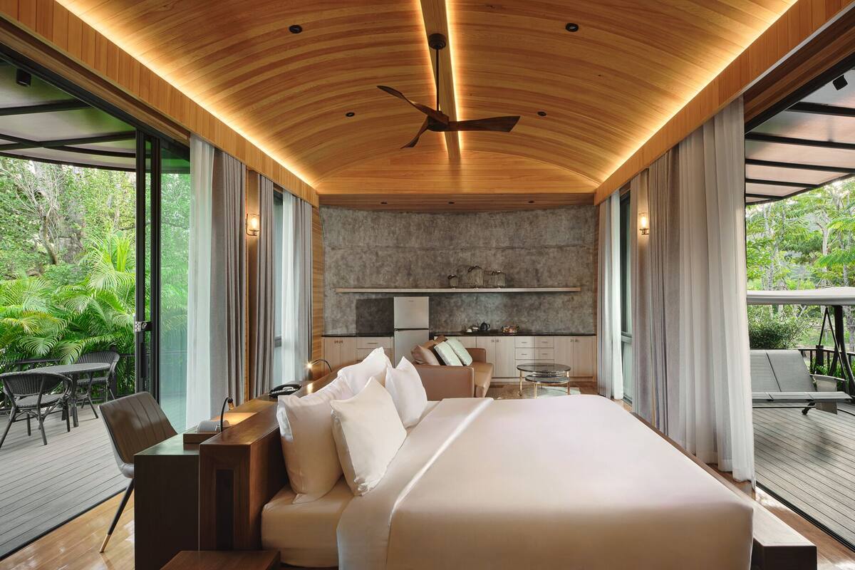 Un dormitorio moderno con una cama grande, techo de madera y vista a vegetación exuberante.