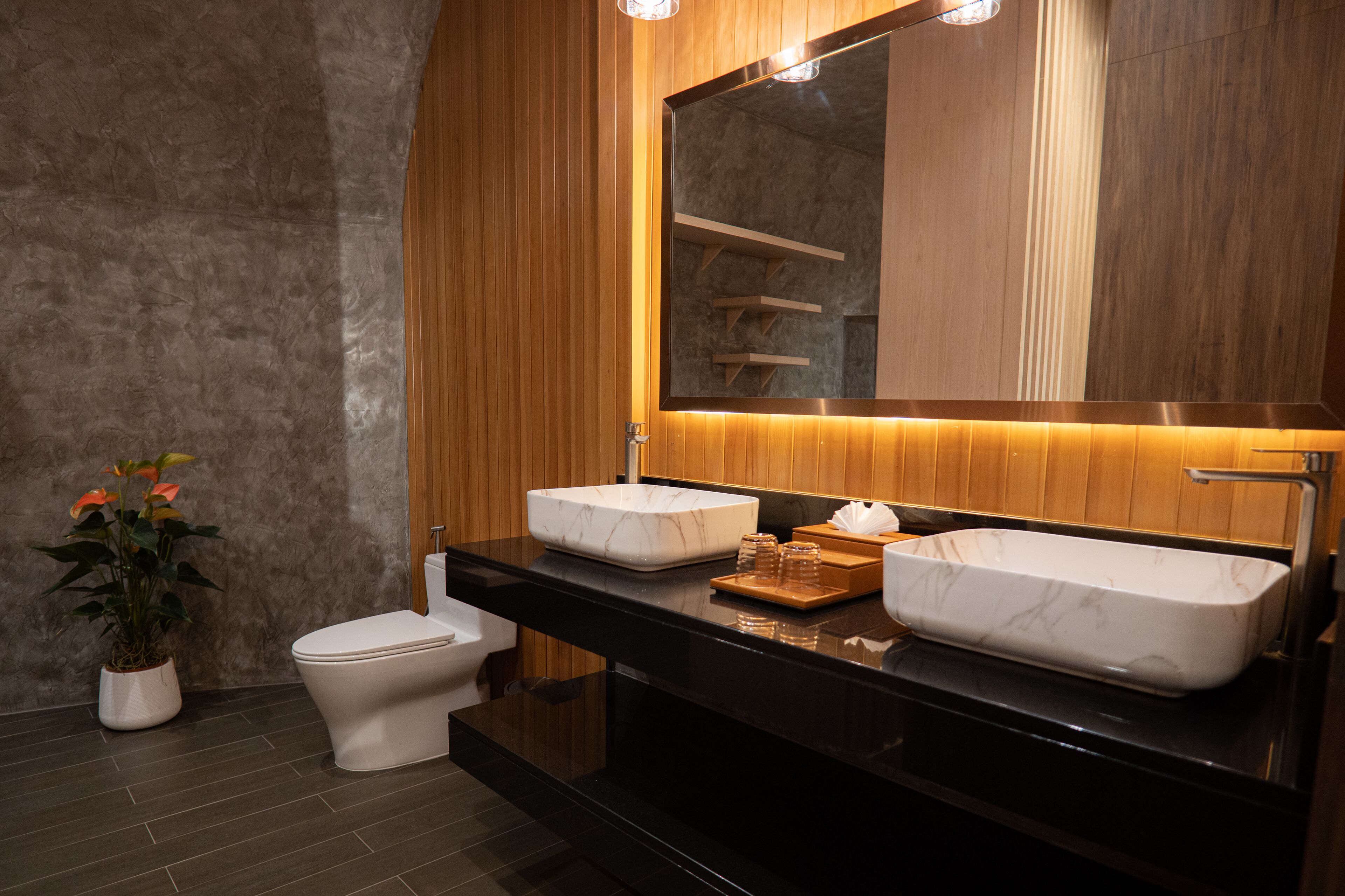 Un baño moderno con tocador de madera, dos lavamanos, inodoro y una planta en maceta.