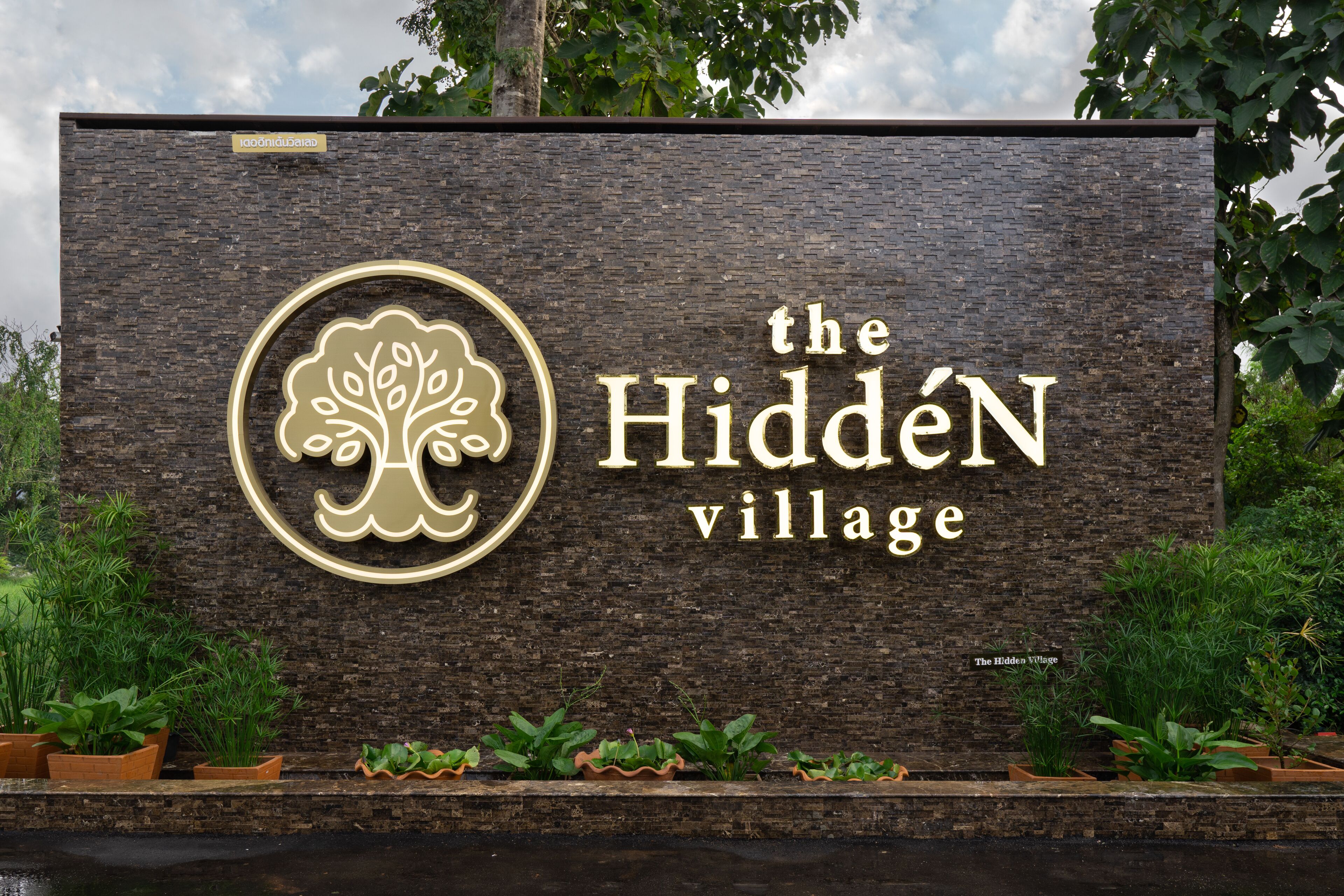A sign for "The Hidden Village" with a tree logo, surrounded by potted plants.
