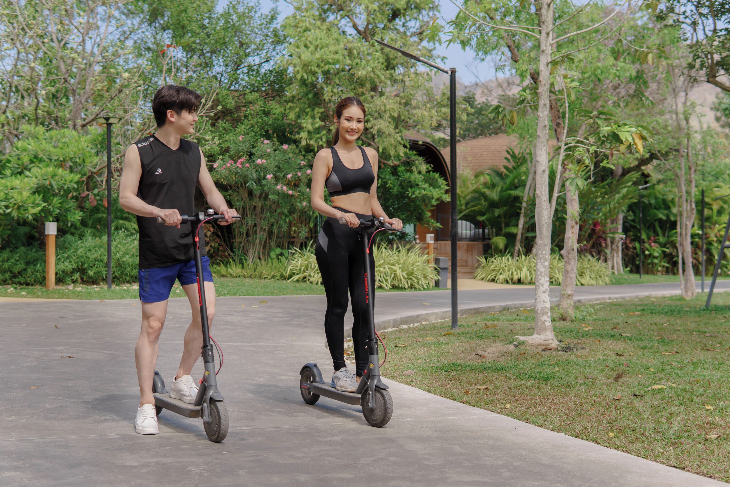 Dos personas montando patinetes eléctricos por un camino pavimentado en un parque con árboles y vegetación.