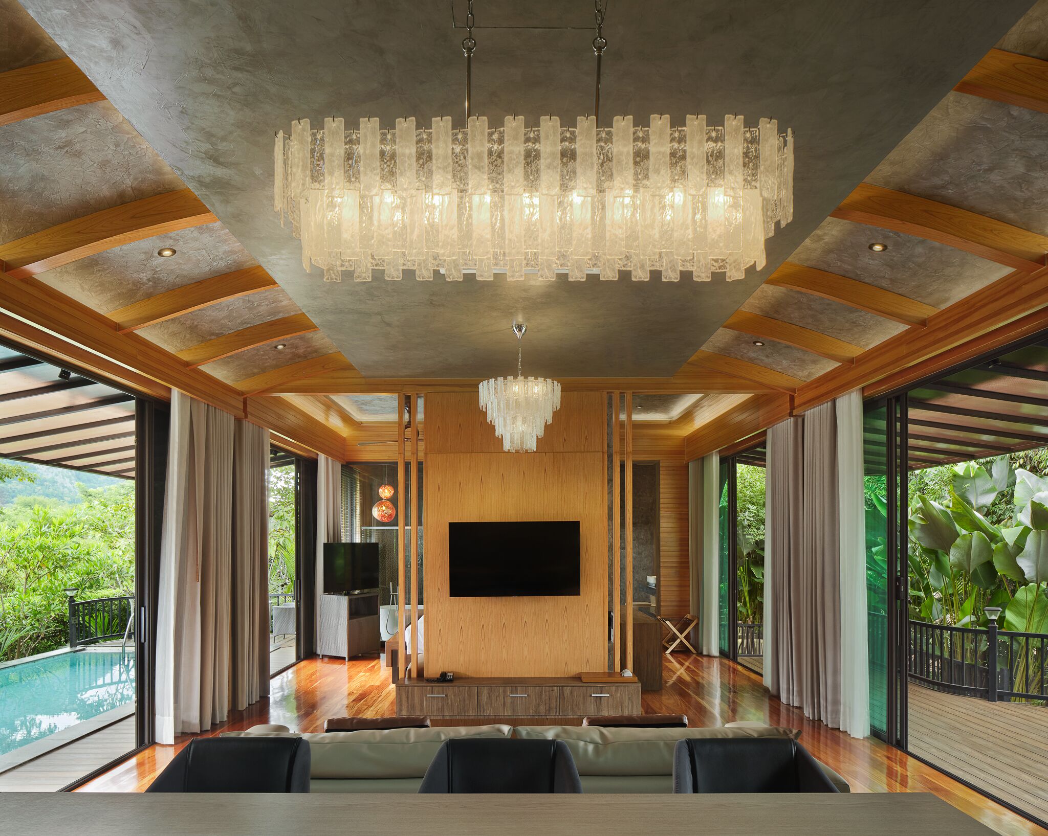 A spacious living room with a large TV, wooden paneling, a chandelier, and a view of a pool and greenery.
