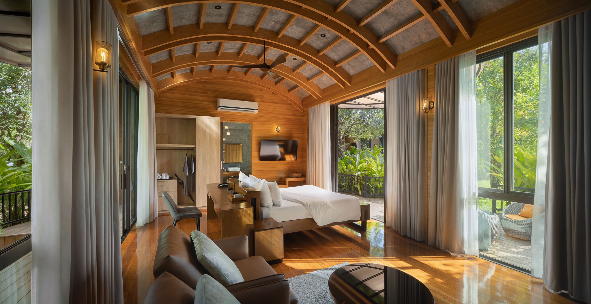 A spacious bedroom with a large bed, a desk, and a seating area. There is a TV mounted on the wall and a ceiling fan. The room has large windows with curtains and a view of greenery outside.