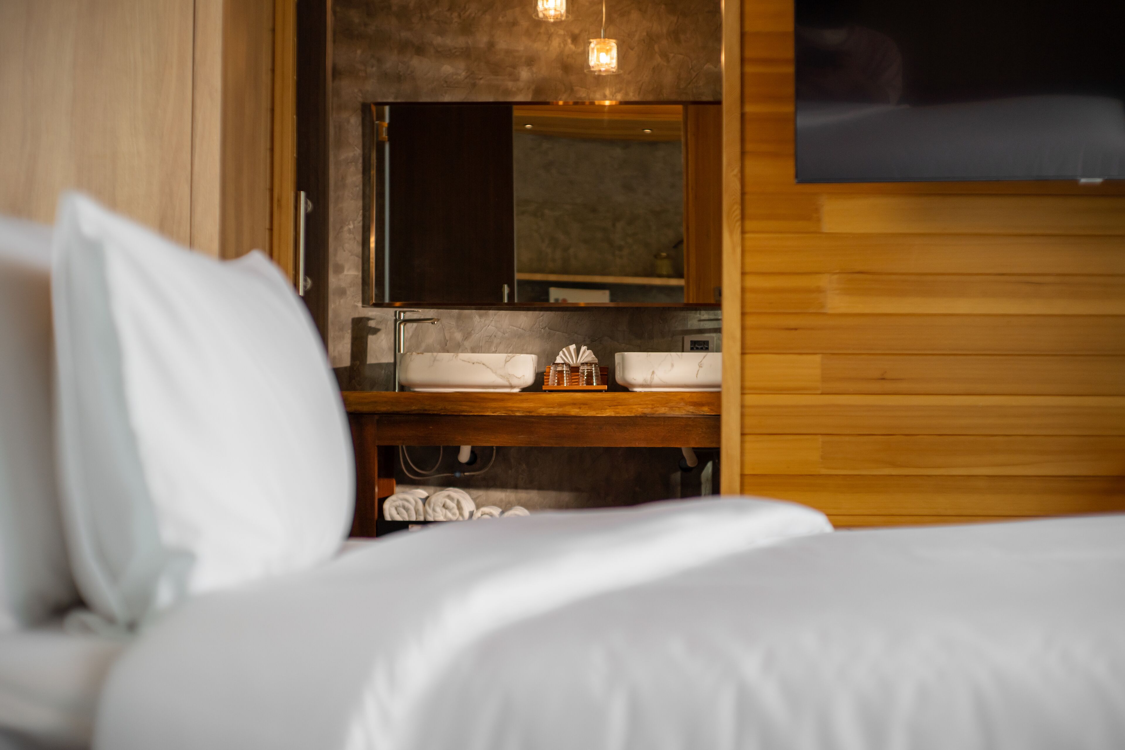 A hotel room with a bed, a wooden headboard, a large mirror, a wooden vanity with a sink, and a television mounted on the wall.