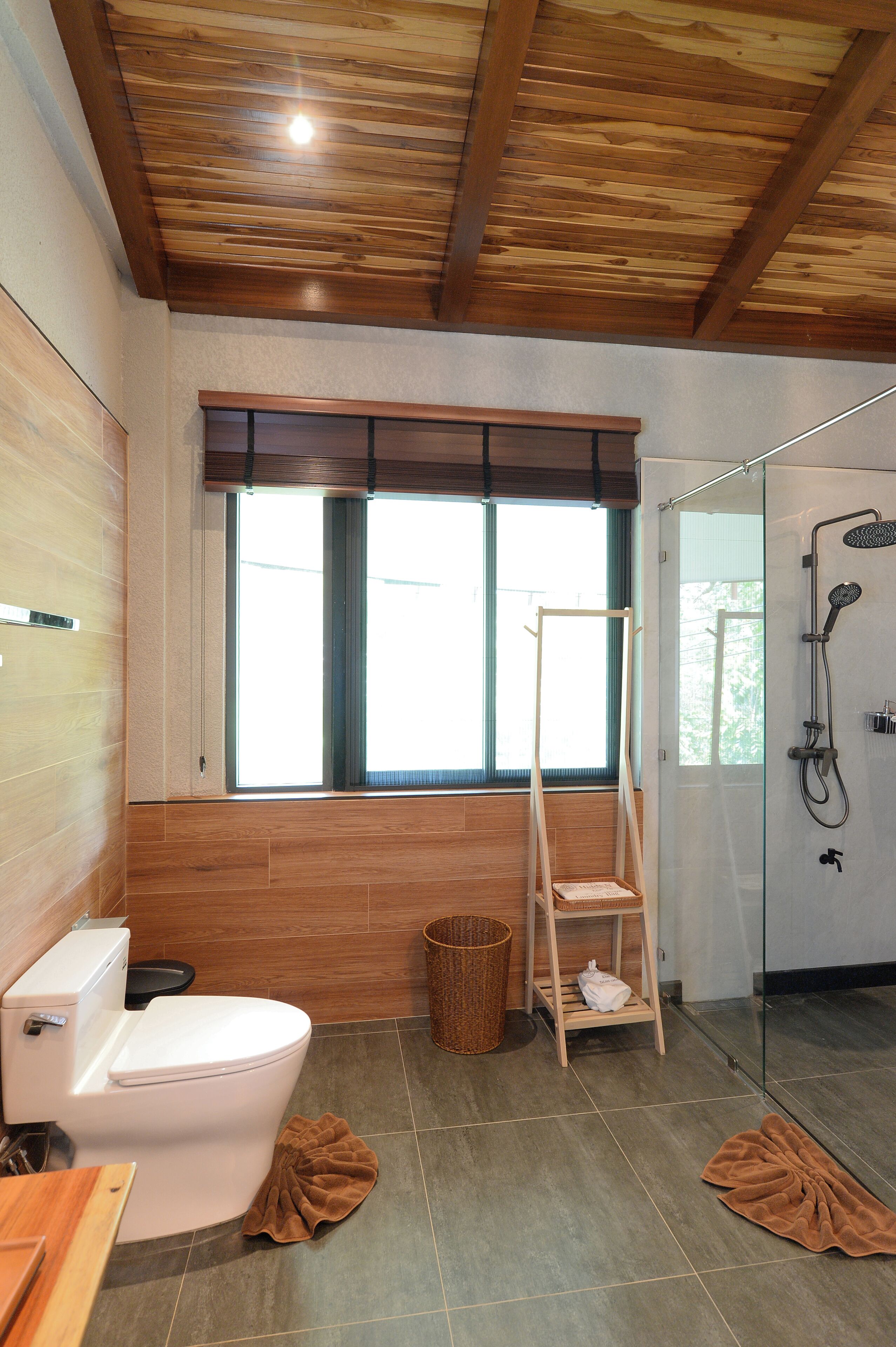 A bathroom with a wooden ceiling, a toilet, a shower, and a window.