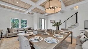 Un salón espacioso con techo de madera, comedor con una mesa larga dispuesta para cuatro personas, y una escalera que sube a un piso superior.