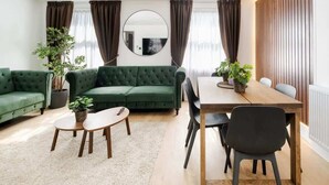 Un salon moderne avec un canapé vert, une table à manger en bois et deux chaises. Un miroir rond est accroché au mur, ainsi qu’une plante en pot et une petite table ronde sur laquelle se trouve une télécommande.