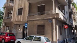 Un immeuble à plusieurs étages doté de balcons, un panneau de signalisation bleu indiquant un parking, et une voiture rouge garée devant.