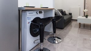 Un espace de vie compact comprenant un lave-linge, un canapé gris et deux tabourets de bar noirs.
