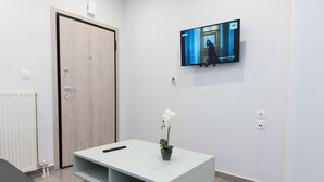 Une chambre d’hôtel moderne avec une porte blanche, un téléviseur fixé au mur, une console blanche sur laquelle reposent une télécommande et un pot d’orchidée, et un radiateur au sol.