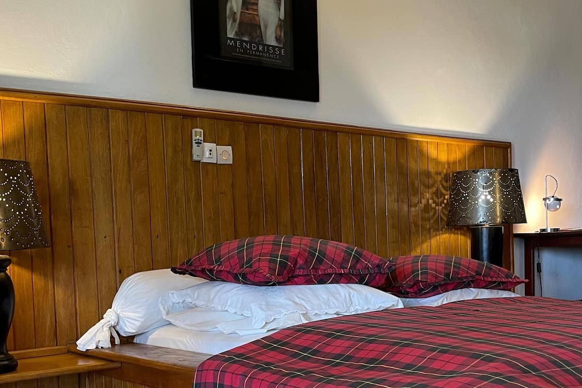 Un dormitorio con cabecera de madera, cama con manta a cuadros rojo y negro, almohadas blancas y dos lámparas de noche.