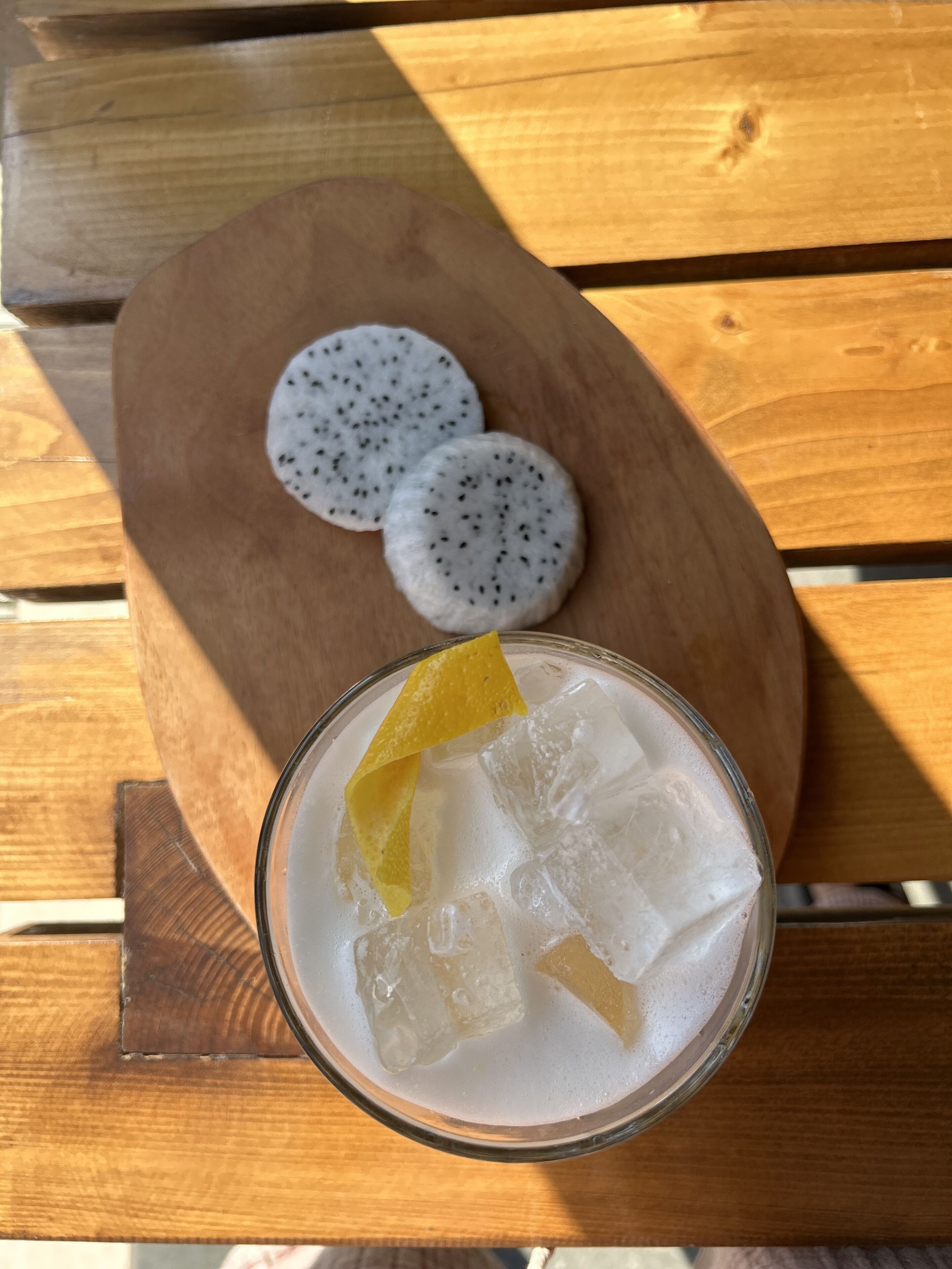 A glass of iced beverage with ice cubes and a slice of fruit, accompanied by two round, white snacks on a wooden board.