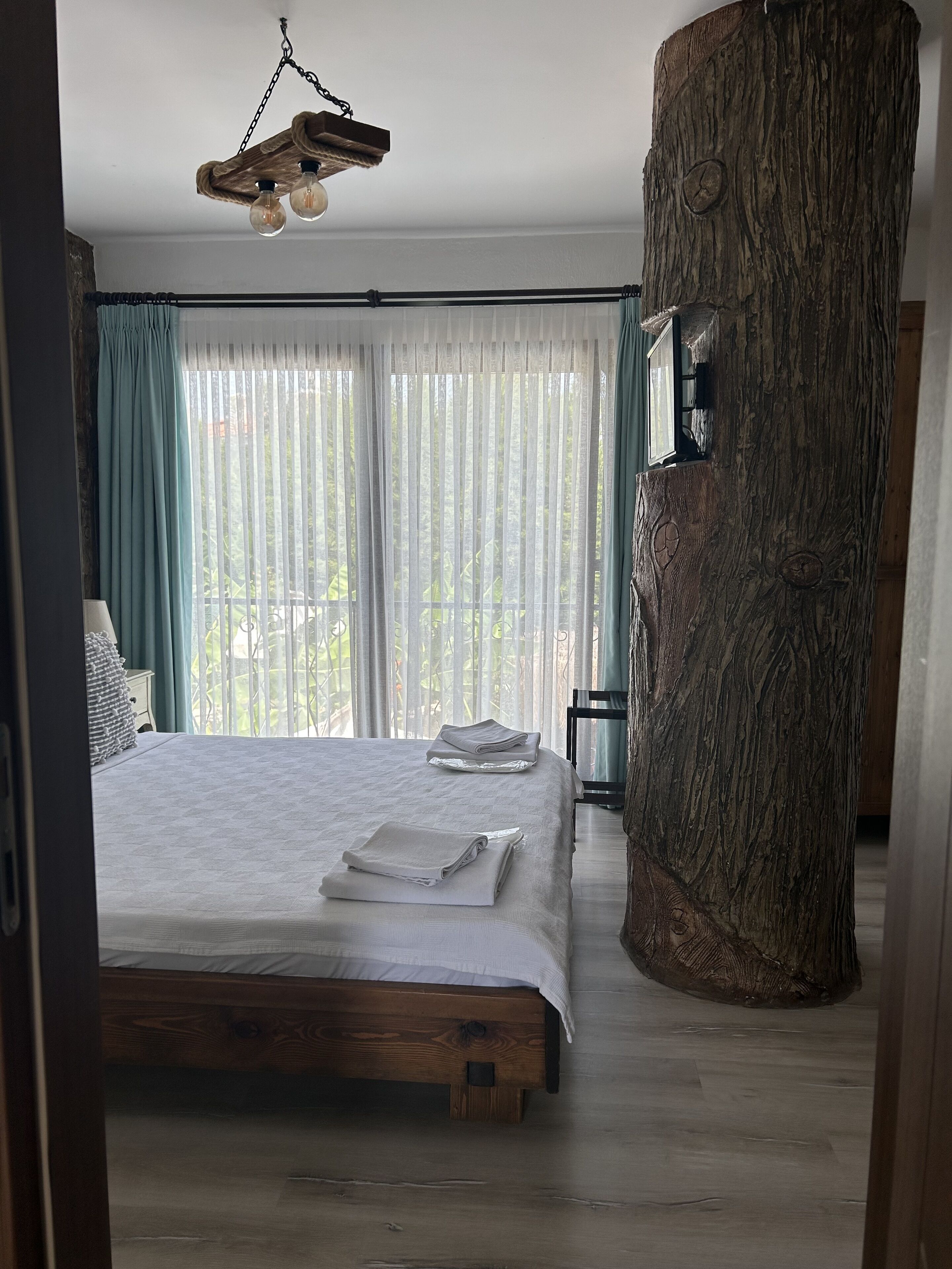 Een slaapkamer met een houten bed, een grote boomstam-zuil, transparante gordijnen en een hanglamp.