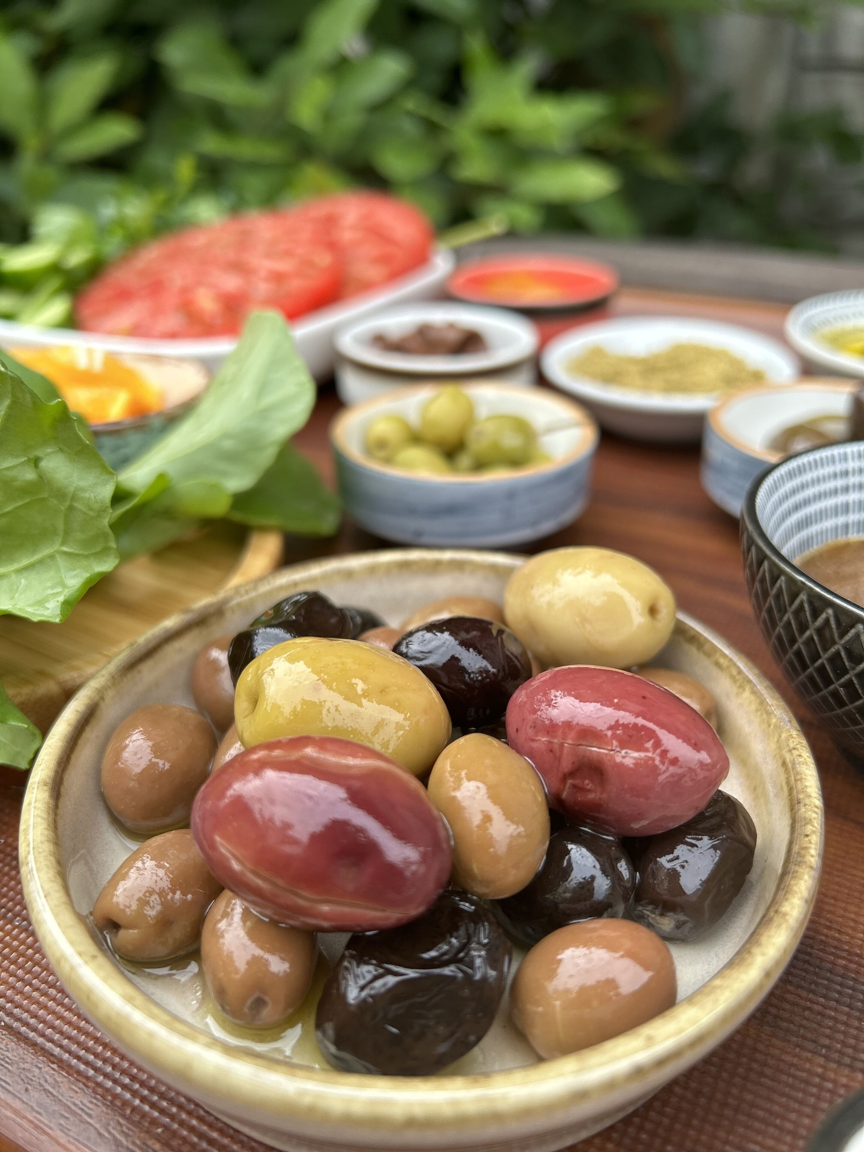 A variety of olives in a bowl, surrounded by other food items and bowls on a table.