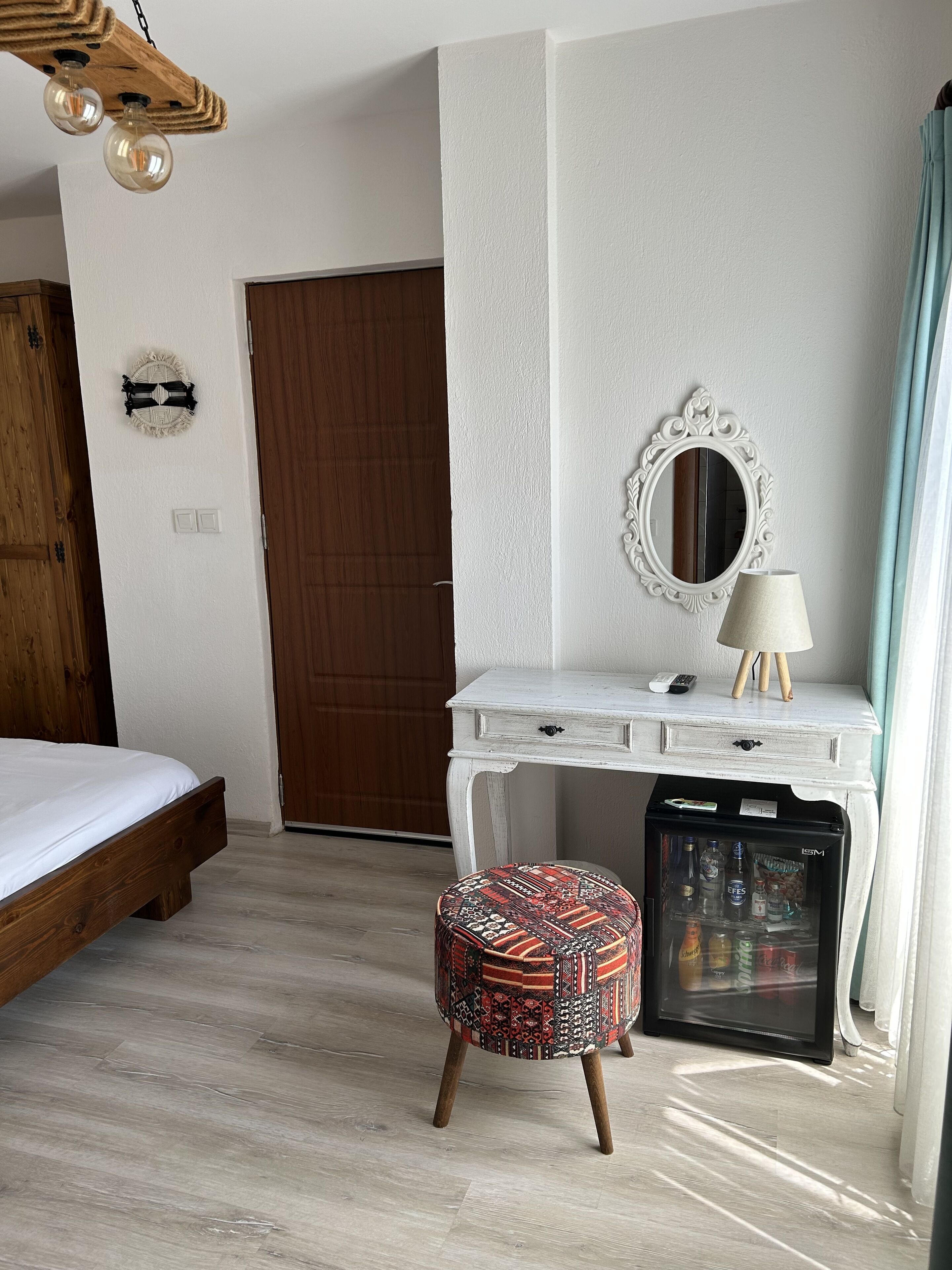 Une chambre d’hôtel avec un lit, une commode blanche, un miroir, une lampe, un petit réfrigérateur et un pouf à motifs.