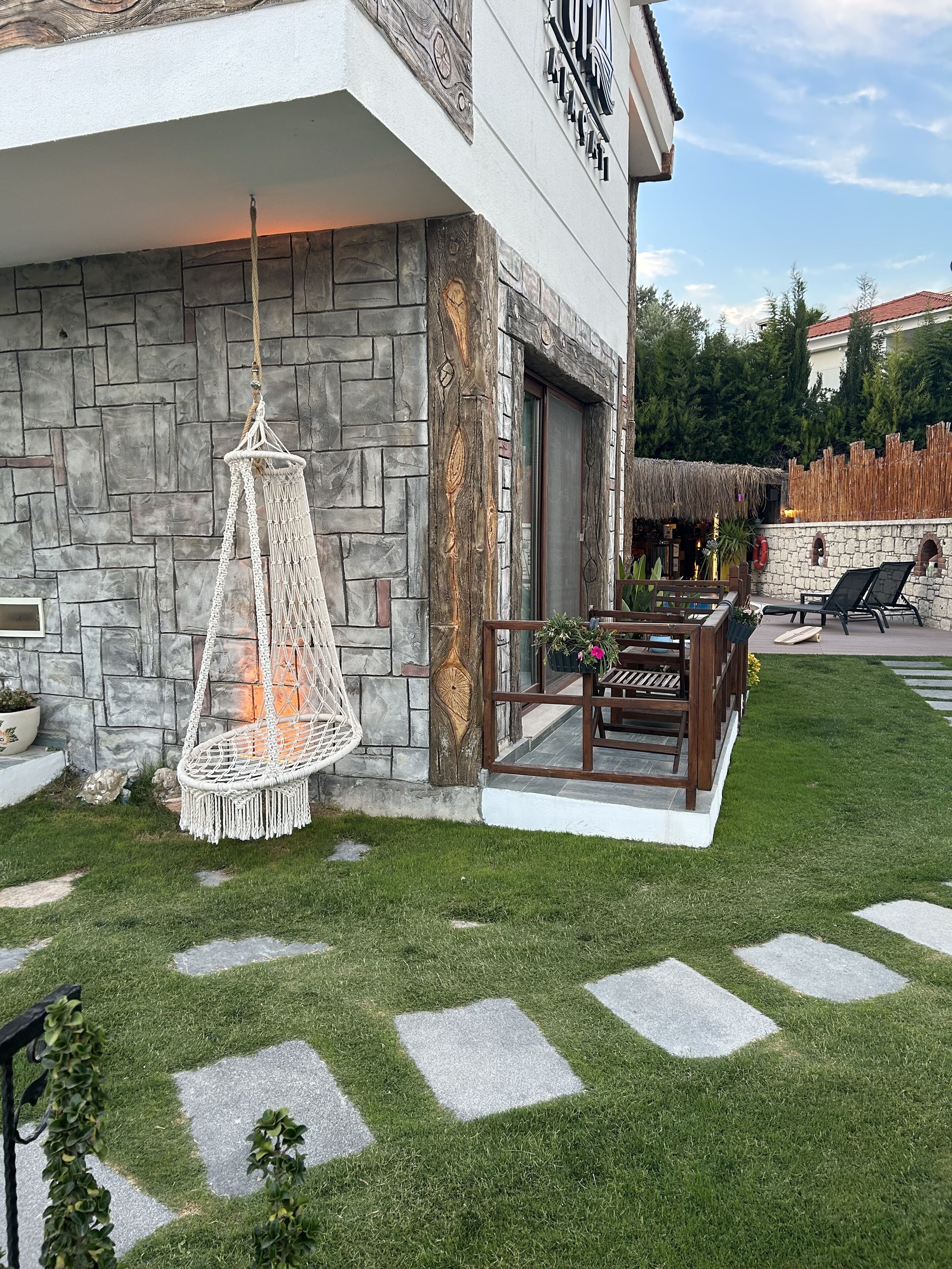 Un bâtiment moderne avec une façade en pierre, un fauteuil suspendu en macramé, des sièges d’extérieur et une structure surmontée d’un toit de chaume.