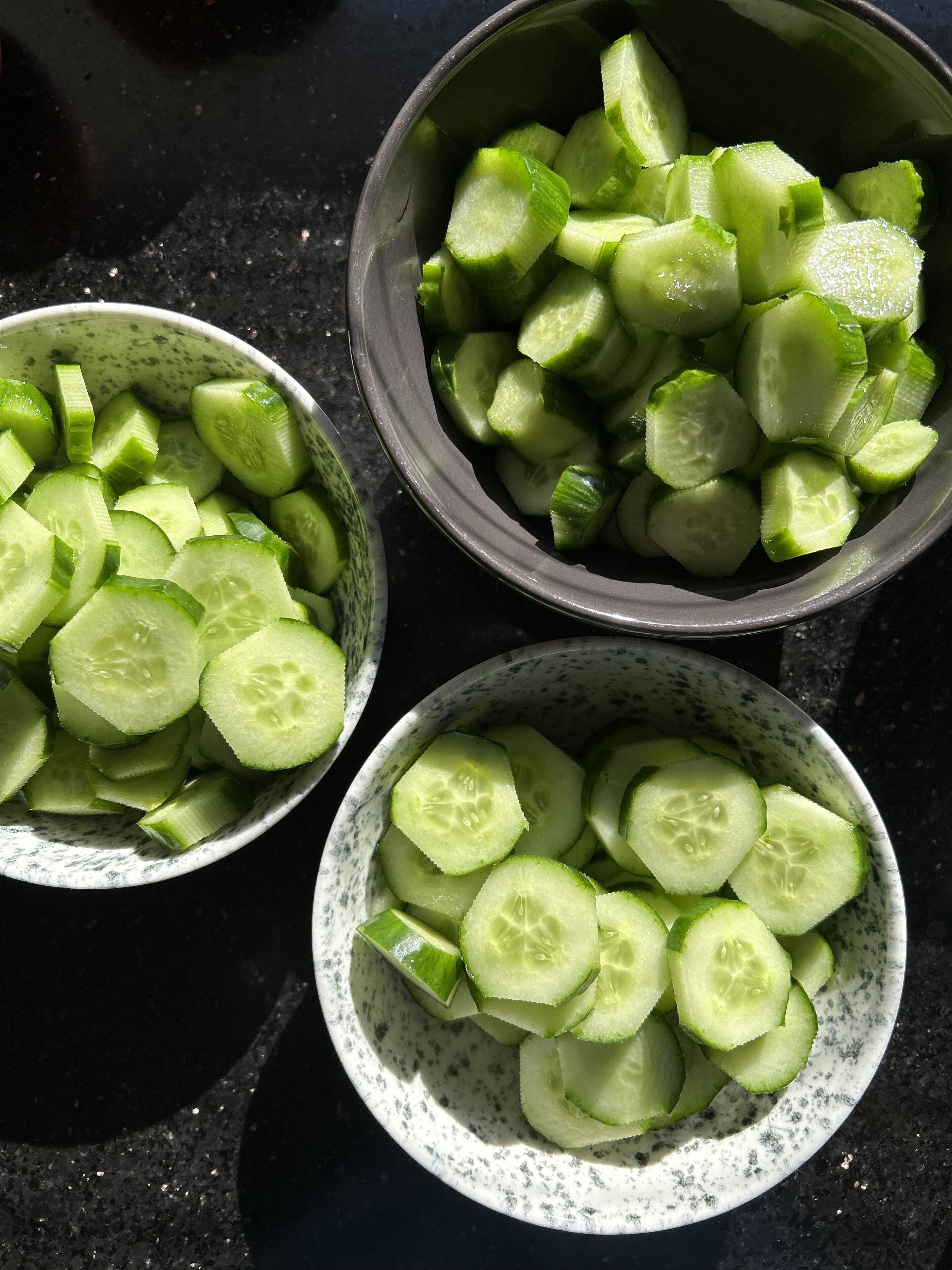 Three bowls of sliced cucumbers on a dark surface.