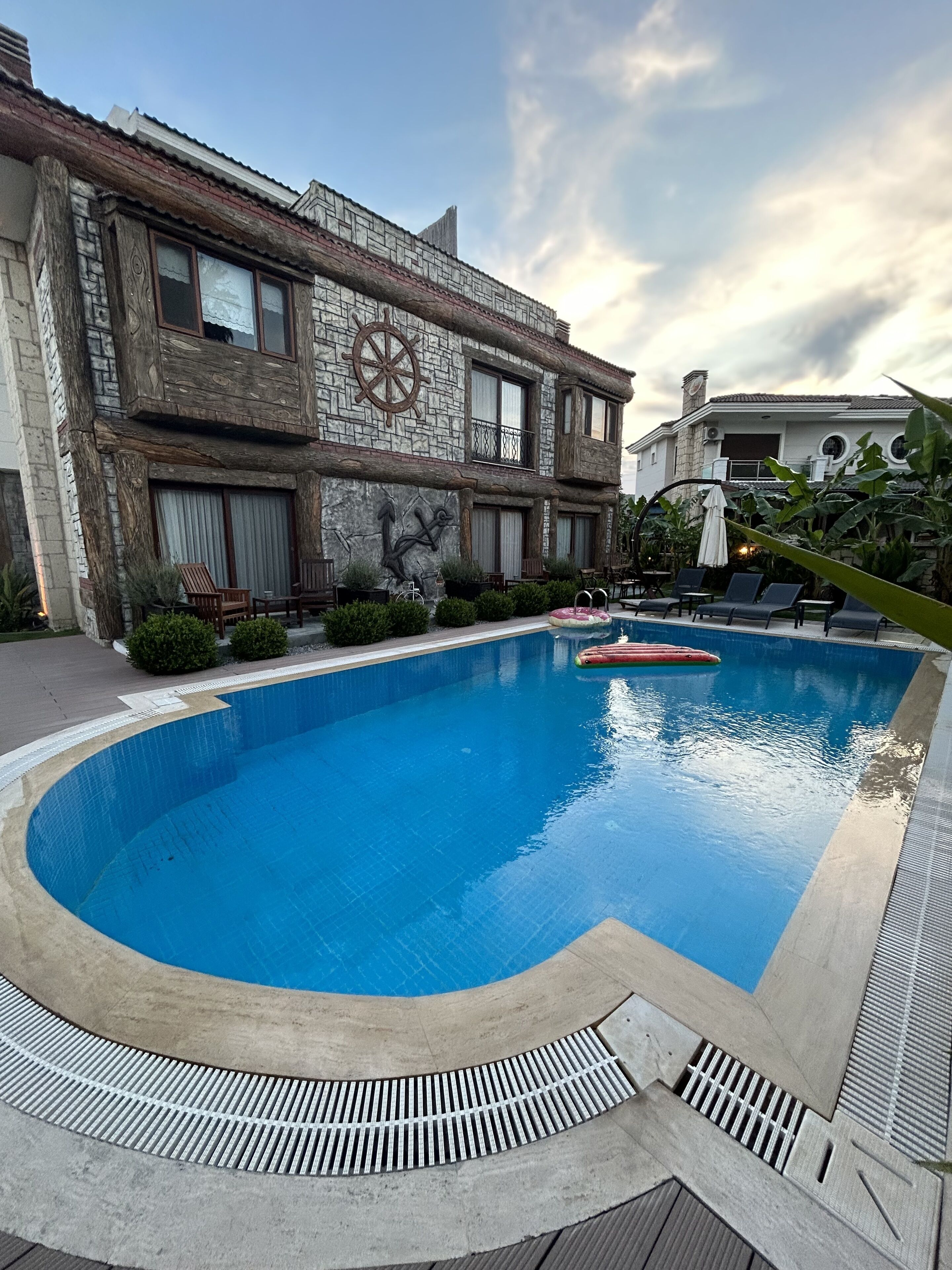 Une piscine dotée d’une façade en pierre et d’une grande fresque représentant une ancre, entourée de chaises longues et d’un jardin bien entretenu.