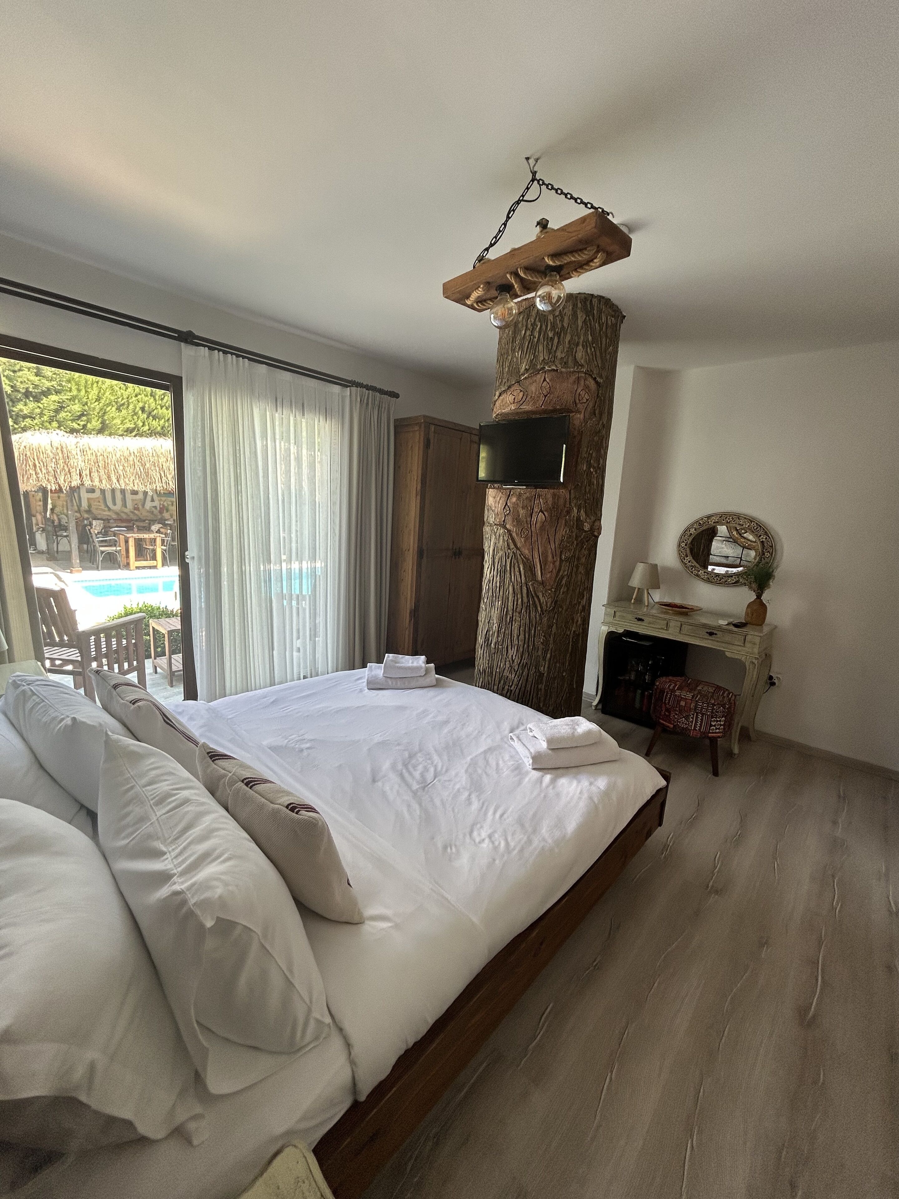 Une chambre spacieuse avec un grand lit, une colonne en bois, une télévision, un bureau avec une lampe et une vue sur l’espace extérieur abritant une piscine.