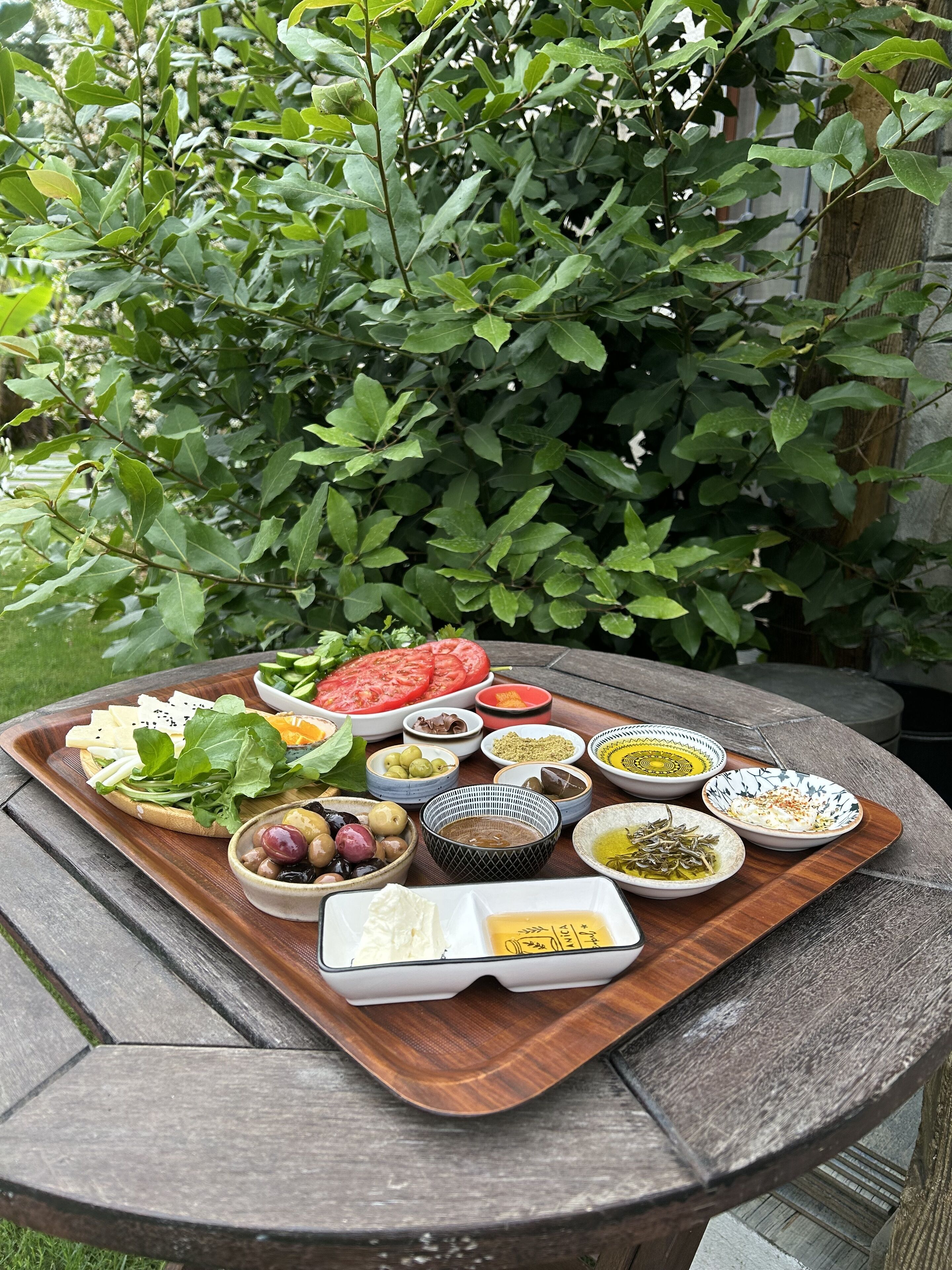 A wooden table with a variety of food items and bowls, set outdoors with a lush green background.