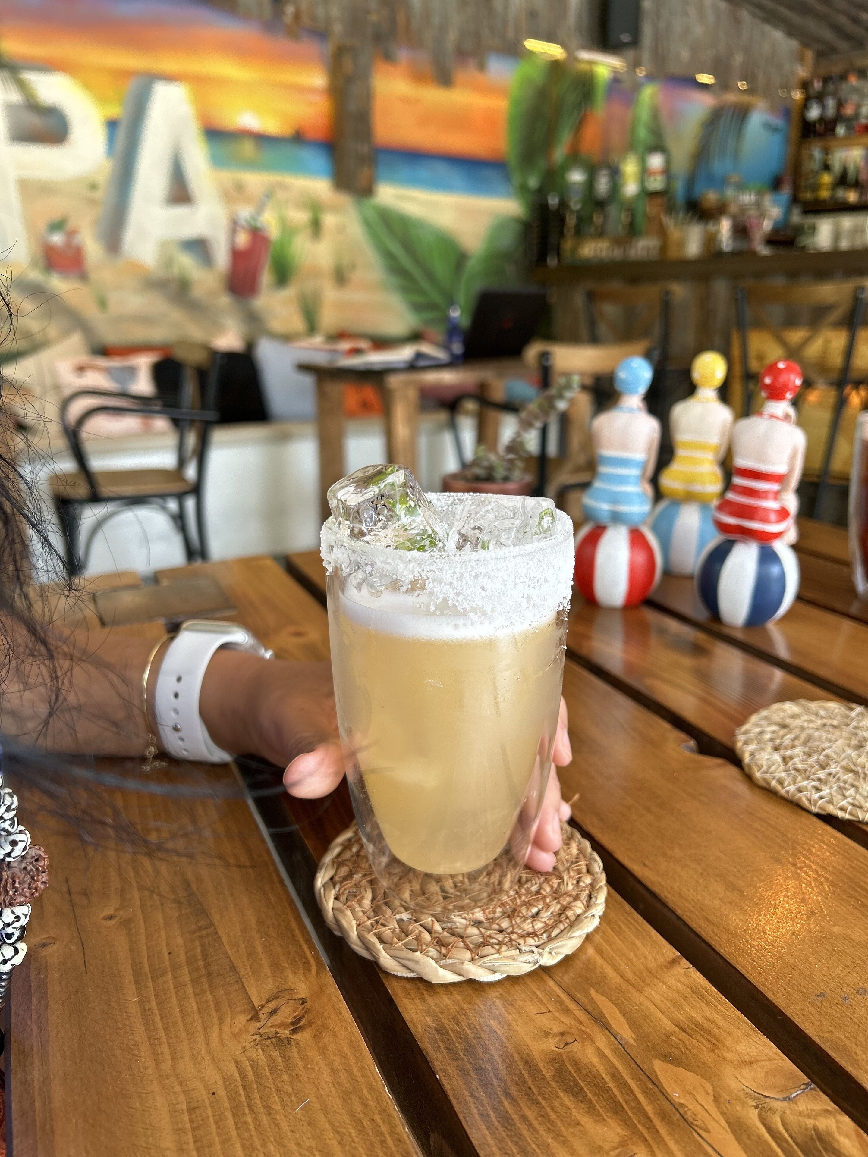 Eine Person hält ein Glas mit einem schaumigen Getränk, im Hintergrund eine Bar mit bunten Flaschen und ein Wandgemälde.