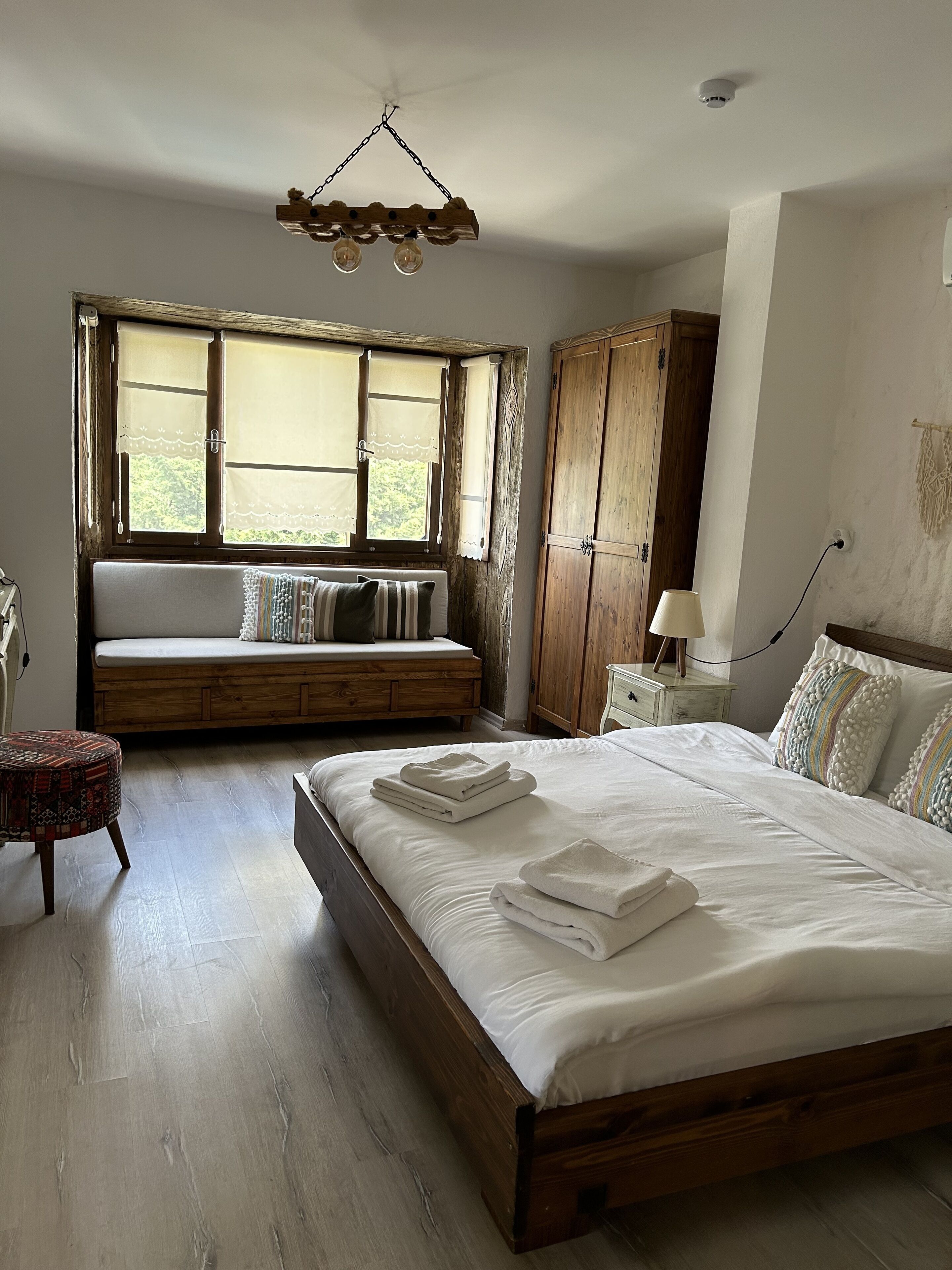 A bedroom with a wooden bed, a bench with cushions, a wooden wardrobe, and a window with curtains.