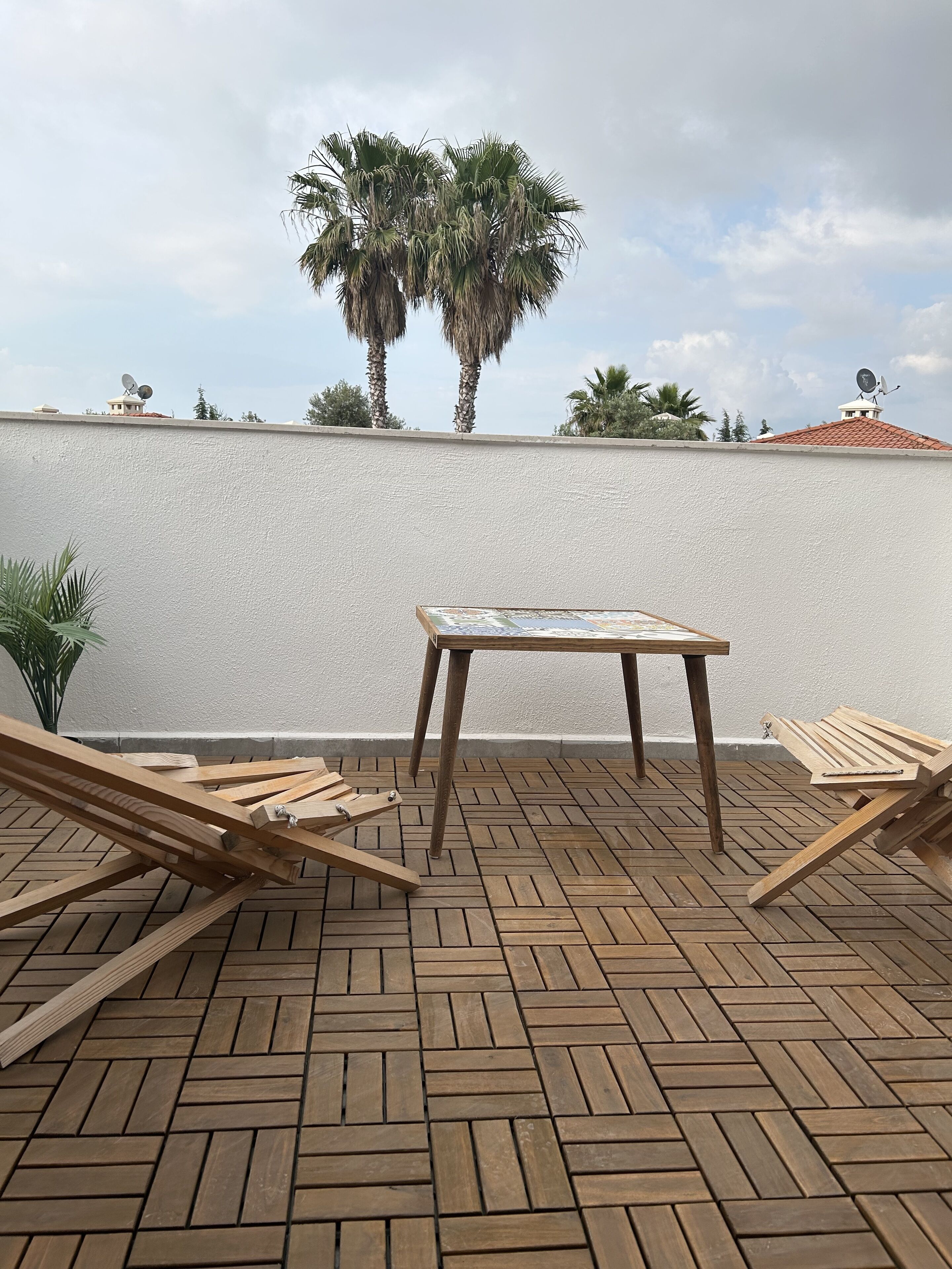 A terrace with a table and two wooden lounge chairs, a white wall, and a palm tree in the background.