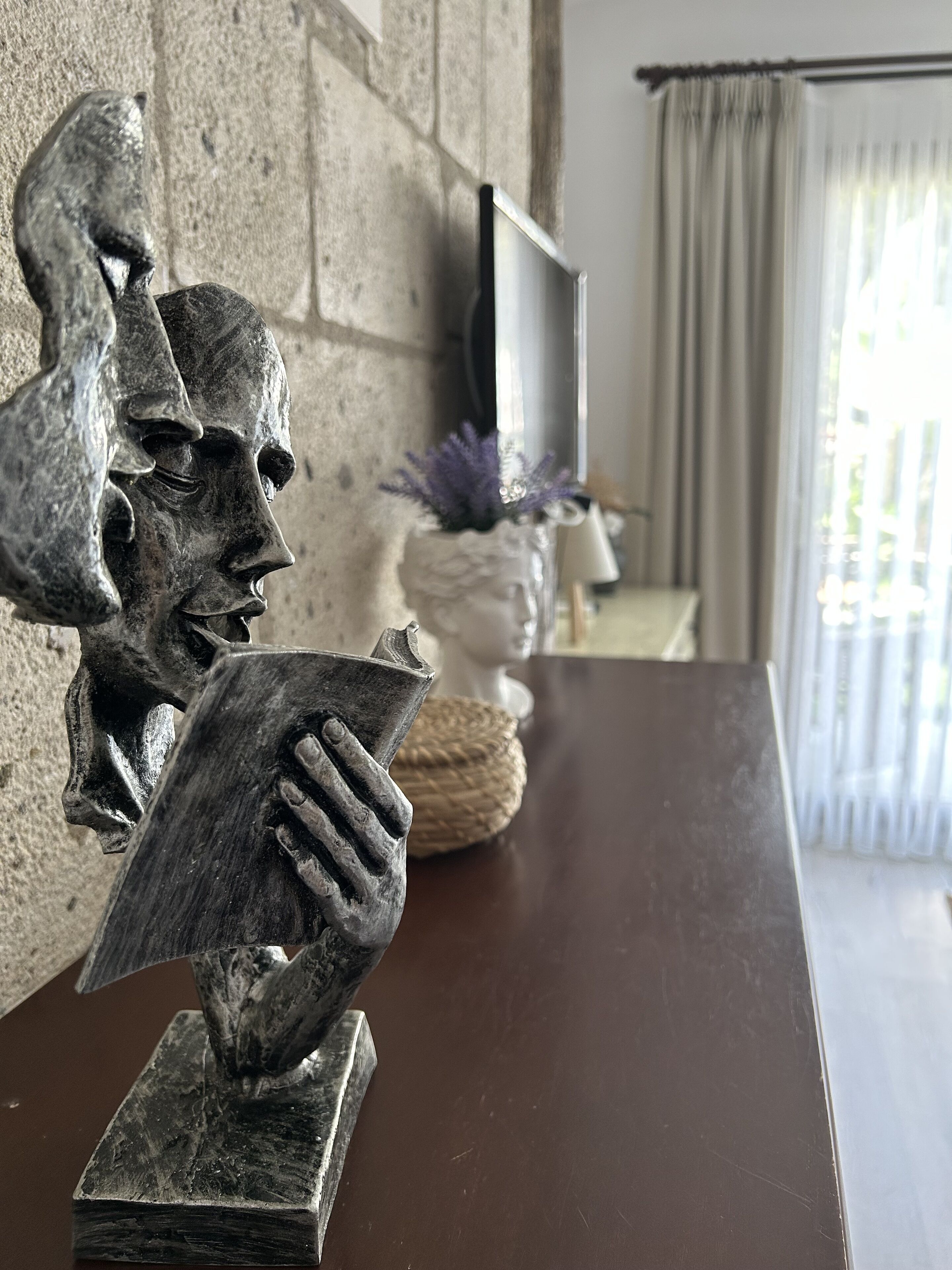A sculpture of a face on a wall, a vase with purple flowers, and a television on a stand.