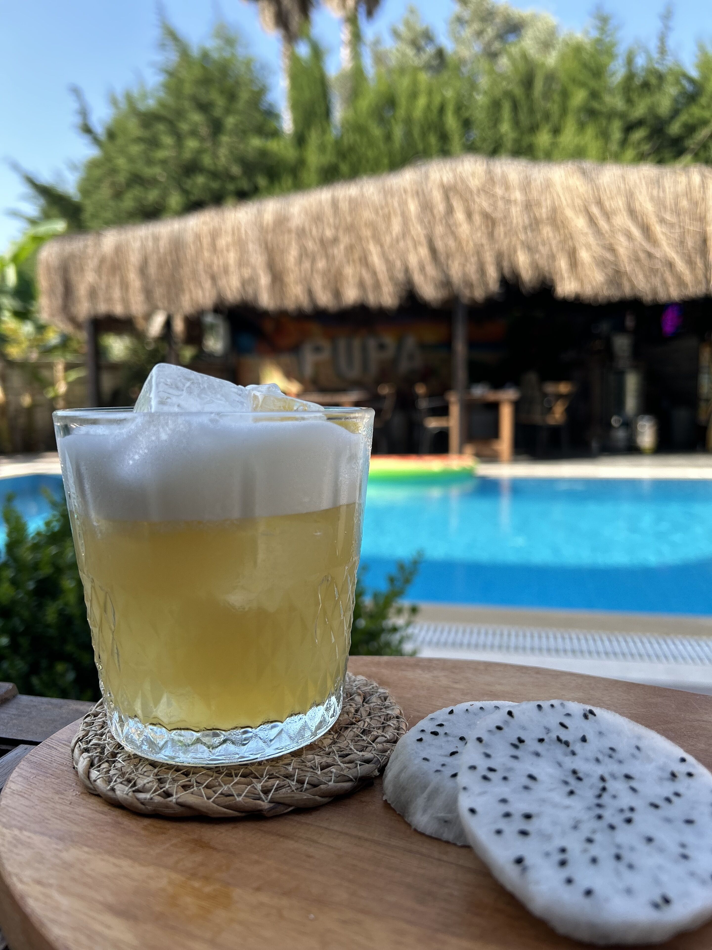 A glass of yellow beverage with ice cubes on a woven coaster, next to a white fruit slice, with a pool and a thatched-roof building in the background.