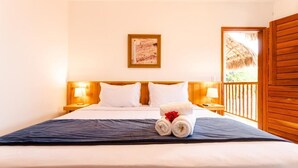 Una cama bien hecha con sábanas blancas, una colcha azul oscuro y dos toallas enrolladas con una flor roja. En la pared sobre la cama hay una obra de arte enmarcada, y se ve una puerta de madera con techo de paja al fondo.
